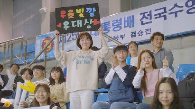 Shin Sol-i holds up a poster for Woo Dae-seong during his swim match which their friends cheer on while sitting. From A Love So Beautiful Kdrama